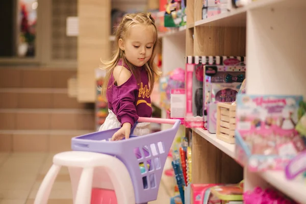 Niña con carrito de compras pequeño en el centro comercial para niños. Chica feliz elegir qué comprar en la tienda de juguetes — Foto de Stock