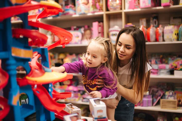 Imádnivaló kislány anyával, aki játékokat vásárol. Aranyos nő a játékboltban. Boldog fiatal lány kiválasztása játék az anyjával — Stock Fotó