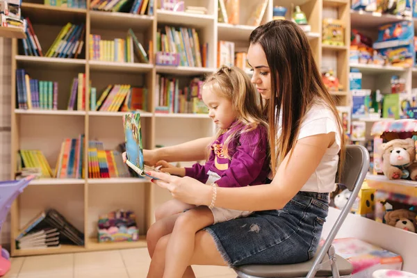 Matka pomoci dcera vybrat interaktivní knihu v hračkářství. maminka a dcera číst knihu v knihovně — Stock fotografie
