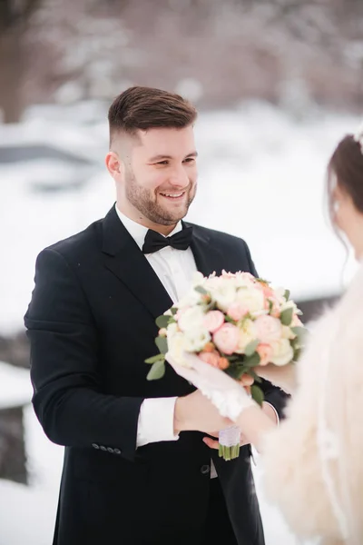 Newlyweds in witer park are walking around. Handsome groom and beautiful bride surrounded by snow. Winter wedding — Stock Photo, Image