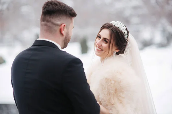 Elegante bruid in witte bontjas staan met knappe bebaarde bruidegom voor besneeuwde bomen — Stockfoto