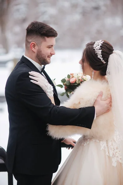 Elegáns menyasszony fehér szőrme kabát állvány jóképű szakállas vőlegény előtt havas fák. — Stock Fotó