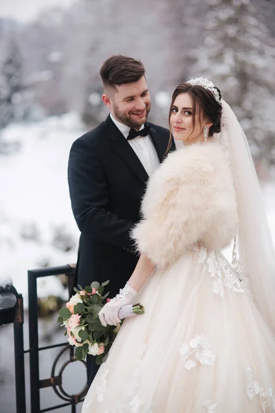 Elegante bruid in witte bontjas staan met knappe bebaarde bruidegom voor besneeuwde dennenbomen — Stockfoto