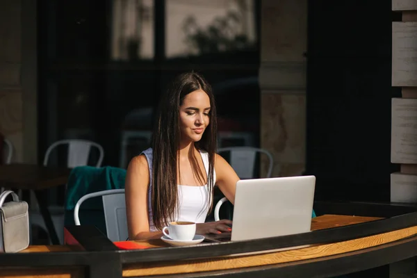 Молодая деловая леди работает на ноутбуке и пьет кофе. Красивая девушка сидит в кафе на террасе. Женщина с телефоном и ноутбуком на открытом воздухе — стоковое фото
