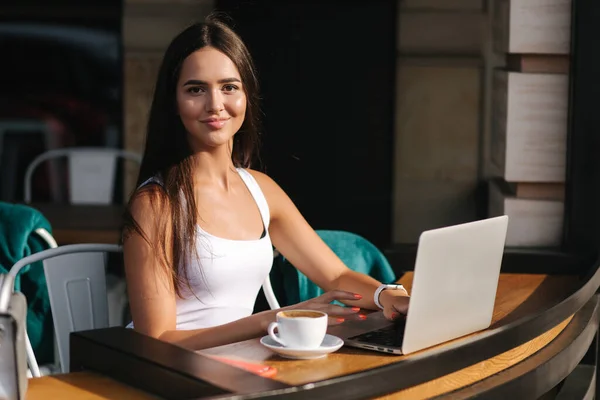 Νεαρή επιχειρηματίας δουλεύει στο λάπτοπ και πίνει καφέ. Όμορφο κορίτσι κάθεται στο καφέ στη βεράντα. Γυναίκα που χρησιμοποιεί τηλέφωνο και φορητό υπολογιστή σε εξωτερικούς χώρους — Φωτογραφία Αρχείου