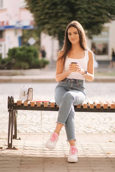 Aantrekkelijke brunette zit alleen op de bank in het centrum van de stad. Mooie jonge vrouw die buiten koffie drinkt. Einde quarantaine. Sociale afstand — Stockfoto