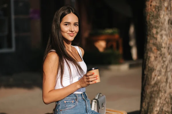 Aantrekkelijke brunette zit alleen op de bank in het centrum van de stad. Mooie jonge vrouw die buiten koffie drinkt. Einde quarantaine. Sociale afstand — Stockfoto