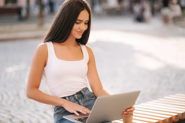 Jovem atraente usar laptop fora no banco no horário de verão. Mulher trabalhando online ao ar livre. Sozinho. Mulher morena bonita. Estudo do estudante — Fotografia de Stock