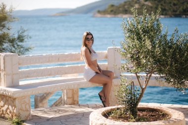 Genç bir kadın güzel bir denizin önünde oturuyor. Suyun mavi rengi. Güneş gözlüklü kadın deniz kenarındaki bankta oturuyor. Yalnız