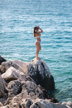 Deniz kenarındaki kayanın üzerinde bikinili seksi kadın. Güzel bir göl. Yaz güneşinin tadını çıkaran kadın. Bikinili seksi bir kadının arka planı