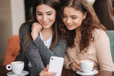 İki kız arkadaşın kafede kahve içip telefonlara baktıklarında. Mutlu, gülümseyen kadınlar terasta oturuyor. İnsanlar hikayelere bakar