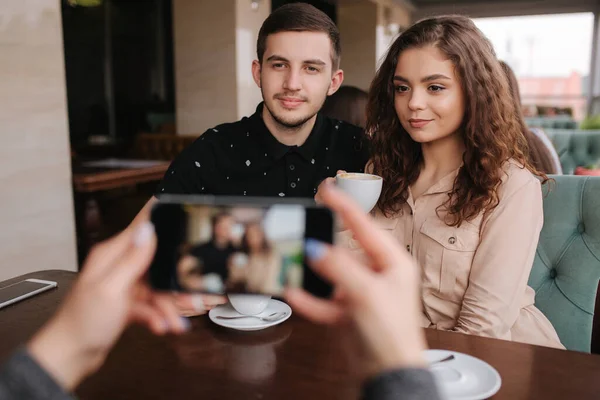 Рука женщины фотографирует красивую молодую пару в кафе. Счастливая улыбающаяся пара позирует для фото на смартфоне. Расслабленный телефон — стоковое фото
