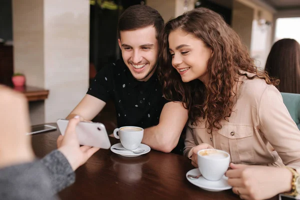 Счастливая пара в кафе смотрит в смартфон и улыбается. Женщина покажет что-нибудь мужчине и женщине по телефону. Человек кладет палец на экран и улыбается — стоковое фото