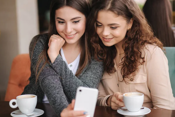 Крупным планом две девушки пьют кофе в кафе и смотрят в Smat телефон. Счастливые улыбающиеся женщины сидят на террасе. Люди смотрят истории — стоковое фото