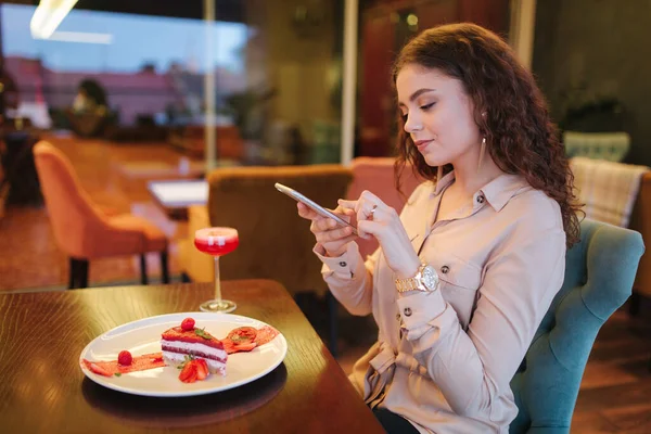 Young woman in restaurant order sweet strawberry cake and red cocktail. Woman make photo of cake