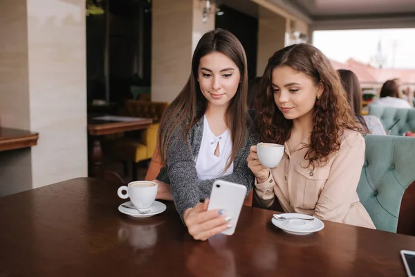 Twee vriendinnen drinken koffie in café en kijken in smat phone. Gelukkig glimlachte vrouwen zittend op het terras. Mensen kijken verhalen — Stockfoto