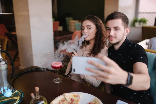 Group of people smoke hookah on terrace. Man and women meet in restaurant. Man make stories while his girlfriend smoke. Man make selfie