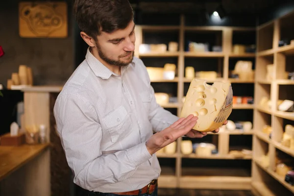 Hansome man hold big slice of cheese maasdam in hand. Cheese with big holes. Background of shelves with cheese