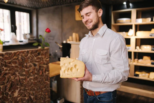 Hansome man houdt een groot stuk kaas maasdam in de hand. Kaas met grote gaten. Achtergrond van planken met kaas — Stockfoto