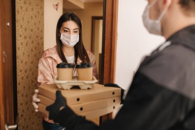 Maskeli ve eldivenli müşterilere pizza ve kahve getiren adam. Kadın kapıyı aç ve siparişi al. Coronavirus teması