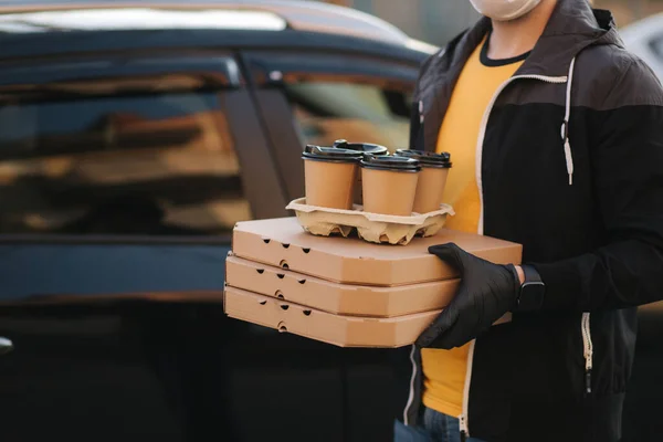 Kurir i skyddsmask och medicinska handskar tar order från bilen. Förlossningspojken håller pizza och kaffe. Leveransservice i karantän. Tema Coronavirus covid-19 — Stockfoto