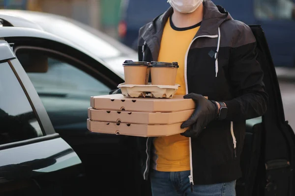 Kurir i skyddsmask och medicinska handskar tar order från bilen. Förlossningspojken håller pizza och kaffe. Leveransservice i karantän. Tema Coronavirus covid-19 — Stockfoto