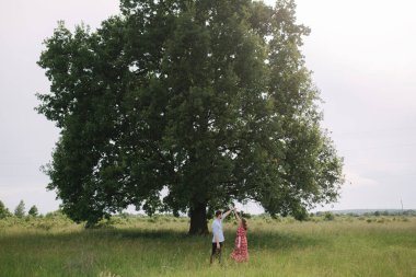 Büyük ağacın önünde yürüyen genç bir çift. Mutlu adam ve kadın birlikte zaman geçiriyorlar. Kırmızı elbiseli kadın.