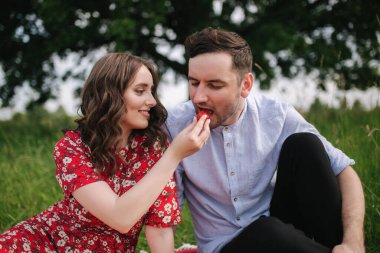 Yakışıklı bir adamla saldıran genç kadın dışarıda kırmızı battaniyenin üzerinde oturuyor. Kadınlar erkeklere çilek verir. Adamın tadı dut gibi