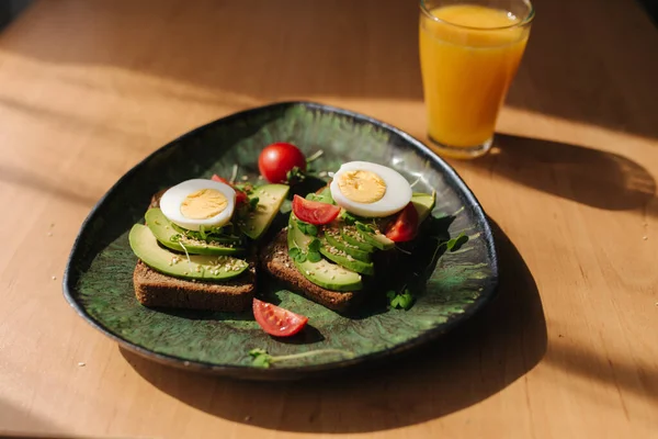 Delicious breakfast at home. Sandwich with fresh sliced avocado above rye toasted bread with cherry tomatoes and boiled egg on green plate. Fresh orange juice — Stock Photo, Image