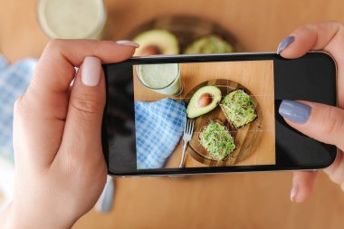 Kadın evde öğle yemeği fotoğrafı çekiyor. Dişi glogger yemek fotoğrafçılığı yapıyor. Sağlıklı vejetaryen yemeği. Avokadolu tost ve ıspanaklı smoothie.