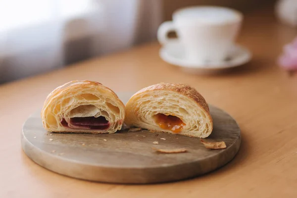 Zwei Hälften frisch entblößtes Croissant auf Holzbrett mit verschiedenen Marmeladen. Hintergrund des Cappuccino — Stockfoto