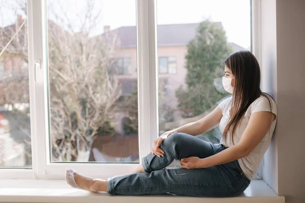 A fiatal nő oldalnézete az ablakpárkányon és védőmaszkban ül, és kinéznek az ablakon. Egyedül otthon a karanténban. Coronavirus téma. Maradj otthon. — Stock Fotó