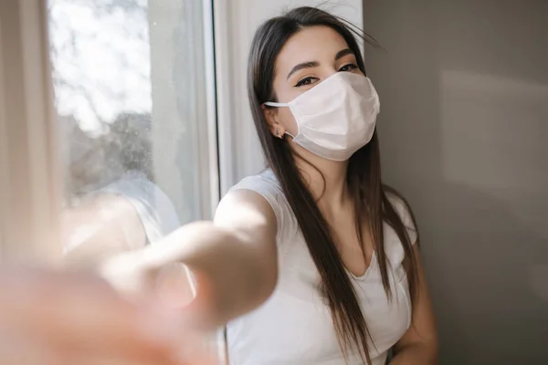 Tıbbi maskeli çekici kadın evde selfie çekiyor. Beyaz tişörtlü ve beyaz maskeli bir kadın telefon kullanıyor. — Stok fotoğraf