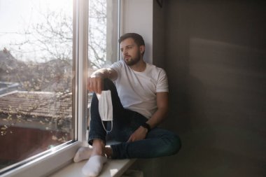 Genç adam pencere pervazına oturur ve ellerini tutar koruyucu maskeyi tutar ve pencereden dışarı bakar. Karantinada evde tek başına. Coronavirus teması. Evde kal.