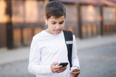 Genç çocuk kablosuz kulaklığı çıkarıp kulaklıklara takıyor. Sırt çantalı beyaz gömlekli genç bir çocuk. Çocuk müziği dinle.