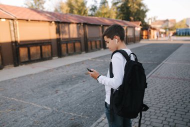 Beyaz tişörtlü, siyah sırt çantalı, telefon, kablosuz kulaklık ve sosyal ağda yazılı mesaj kullanan genç bir çocuk.