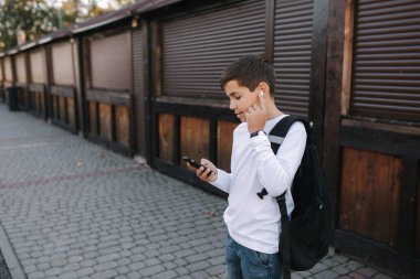 Genç çocuk kablosuz kulaklığı çıkarıp kulaklıklara takıyor. Sırt çantalı beyaz gömlekli genç bir çocuk. Çocuk müziği dinle.