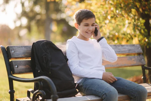 Jonge jongen in wit sweatshirt met zwarte rugzak zittend op de bank in het park en spreek met iemand aan de telefoon — Stockfoto