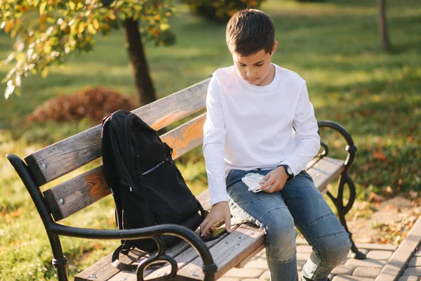 Pohledný teenager si vezme z batohu štěkot za nabíjení chytrého telefonu. Roztomilý chlapec v parku v podzimním čase — Stock fotografie