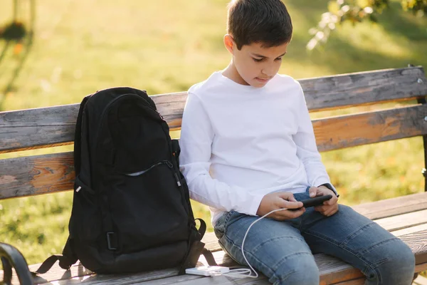 Jovem menino jogar jogos online no smartphone e usando powerbank para carregar. Adolescente bonito usar telefone otside — Fotografia de Stock