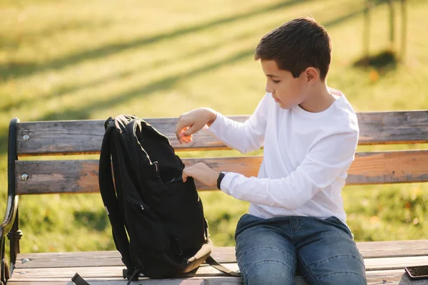 英俊的少年因为给智能手机充电而从他的背包动力树皮中取出。 秋天里公园里可爱的男孩 — 图库照片