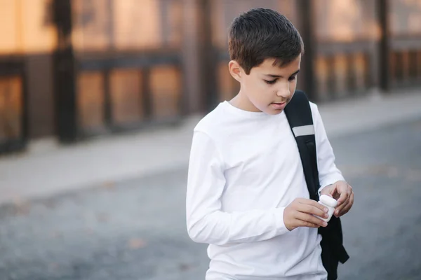 Tienerjongen pakt een draadloze koptelefoon en stopt die in de oren. Jonge jongen in wit shirt met rugzak. Jongen luister muziek — Stockfoto