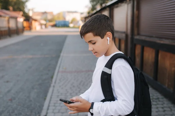 白のスウェットシャツと黒のバックパック付きの10代の男の子は、ソーシャルネットワークで携帯電話やワイヤレスヘッドフォンや印刷メッセージを使用しています — ストック写真