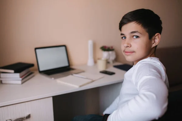 幸せな10代の男の子がテーブルに座って家で勉強します。隔離中にオンラインで勉強する少年。ノートパソコンと本の背景 — ストック写真
