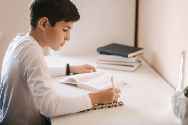 Jonge jongen aan het bureau las het boek en schreef het op in het notitieboekje. Studie thuis tijdens quarantaine — Stockfoto