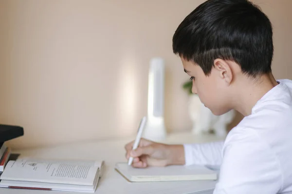 Ein kleiner Junge, der am Schreibtisch sitzt, liest das Buch und schreibt es in ein Notizbuch. Während der Quarantäne zu Hause studieren — Stockfoto