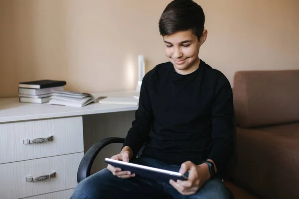 O adolescente em casa senta-se à mesa e usa o comprimido durante o intervalo. Jovem menino jogar jogos online no tablet. Backgroung de mesa com livros — Fotografia de Stock