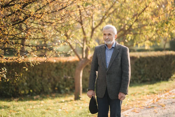 Un bell'uomo anziano che indossa una giacca grigia e un cappello nero. Vecchio barbuto dai capelli grigi passeggia nel parco autunnale. sfondo giallo — Foto Stock