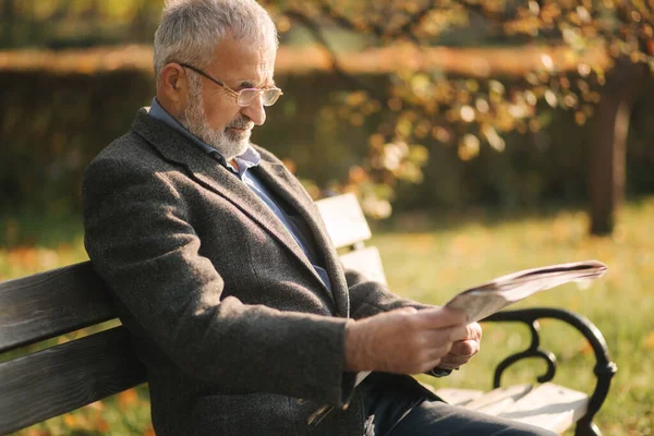 Il nonno bello con una bella barba in giacca grigia si siede su una panchina nel parco e legge un giornale. Uomo anziano dai capelli grigi in occhiali — Foto Stock
