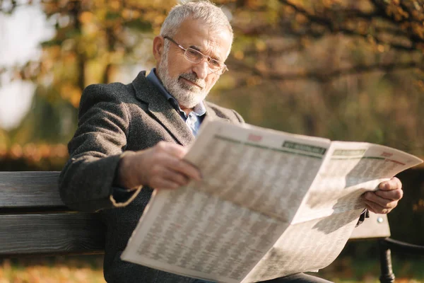 Όμορφος παππούς με ωραία γενειάδα με γκρι σακάκι κάθεται σε ένα παγκάκι στο πάρκο και διαβάζει εφημερίδα. Μεγαλόσωμος γκριζομάλλης με γυαλιά. — Φωτογραφία Αρχείου